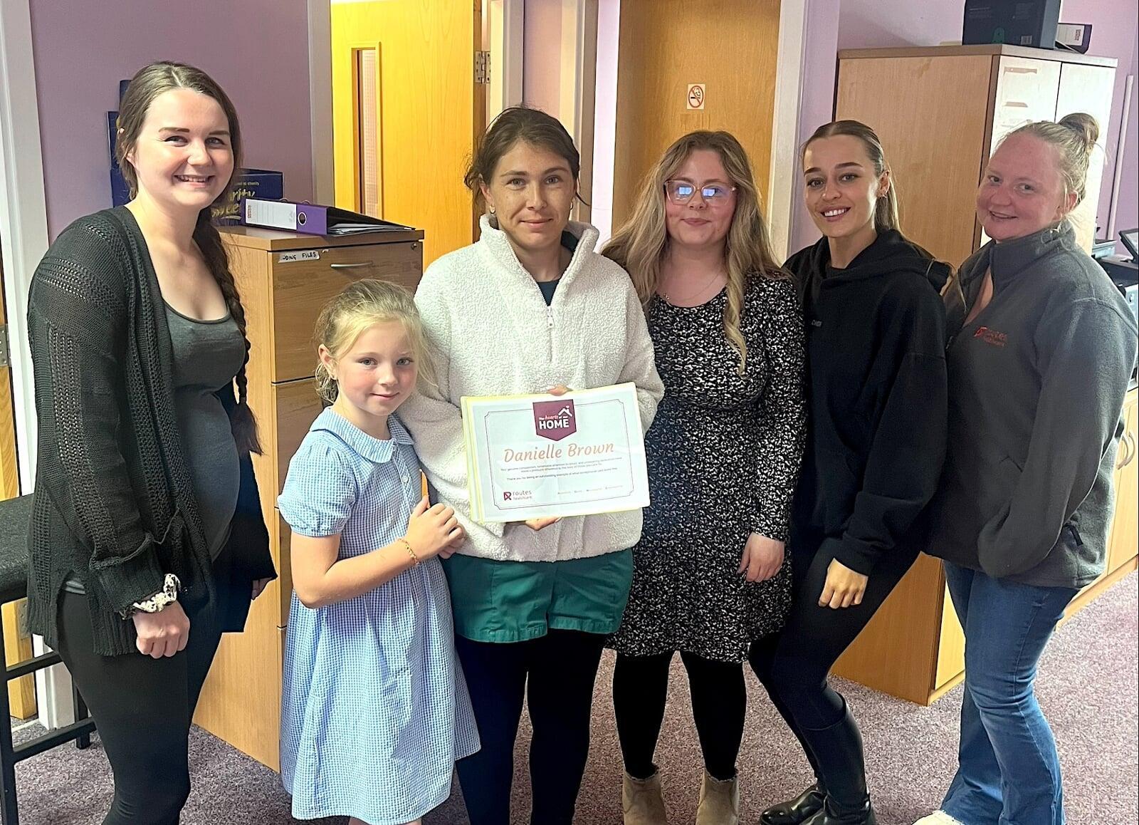 Routes Healthcare Blackpool team celebrating around Danielle Brown's Hearts of our HOME award presentation in office setting